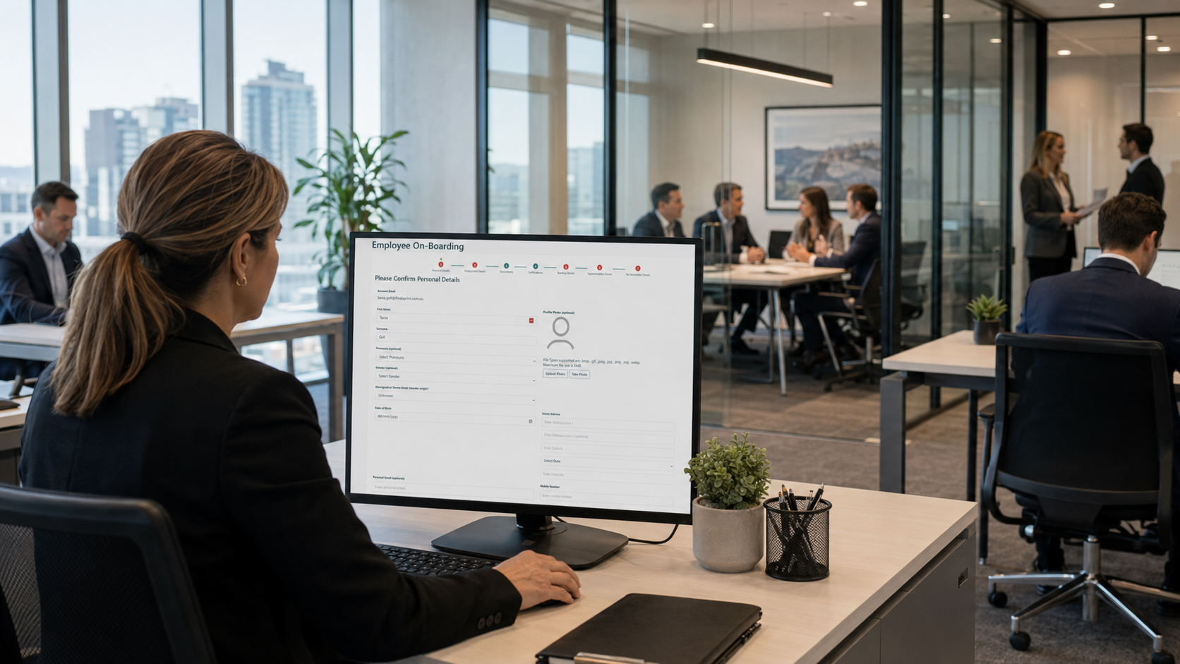 A practice manager using a digital employee onboarding system at her desk while colleagues work and meet in a modern professional services office.